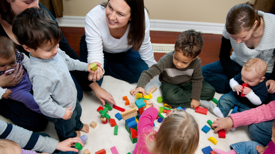 Flera vuxna sitter på golvet med barn i olika åldrar, barnen leker med klossar.