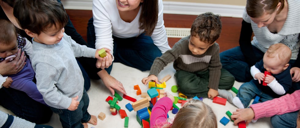 Flera vuxna sitter på golvet med barn i olika åldrar, barnen leker med klossar.