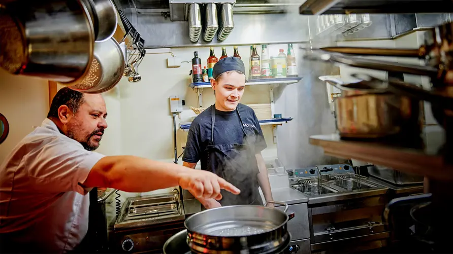 Restaurangkök med en köksmästare och en elev bland stekbord och rykande grytor
