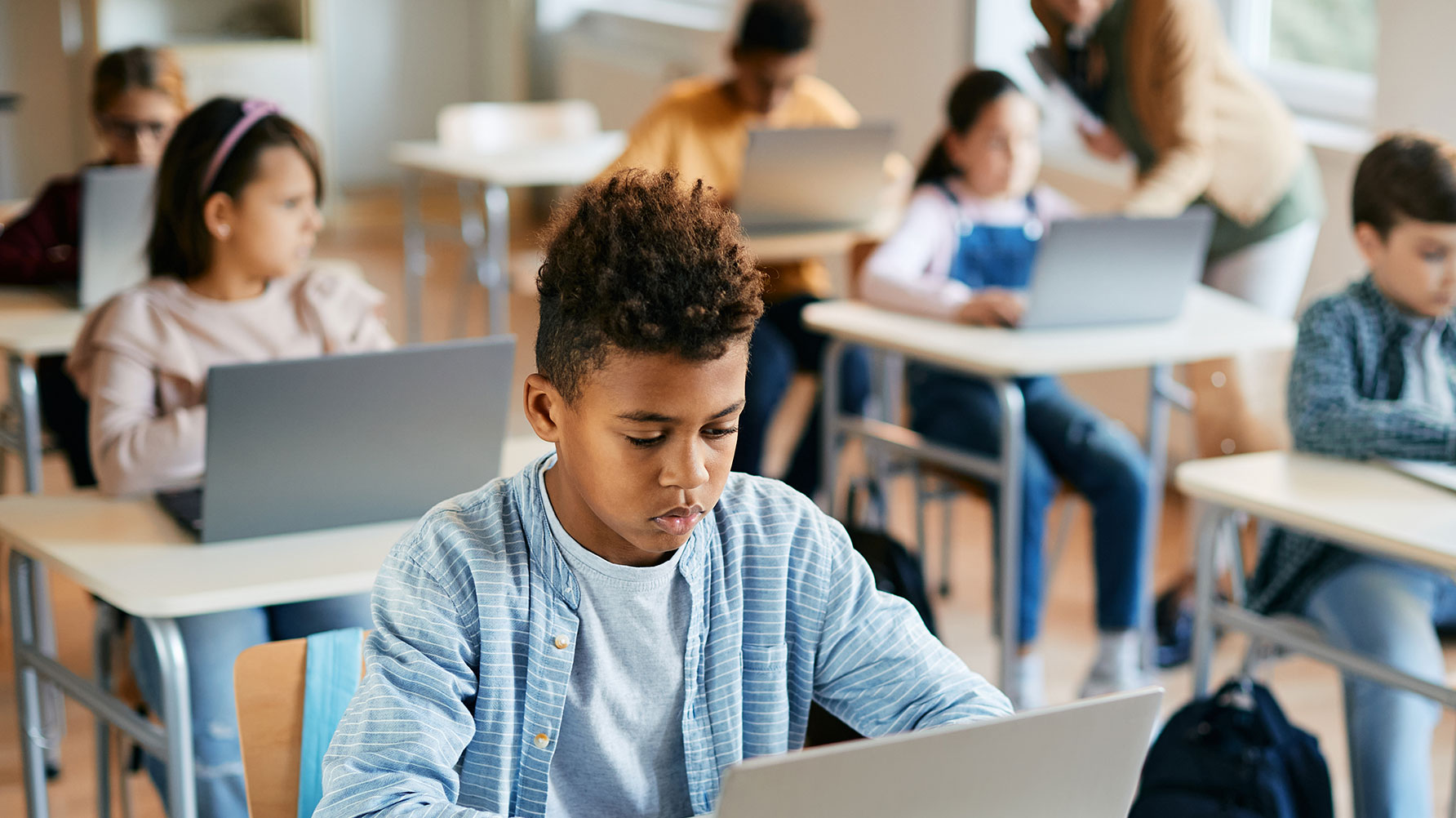 En genrebild med elever som arbetar vid varsin sk&#xE4;rm, eleven n&#xE4;rmast kameran med fokuserat uttryck i ansiktet. I bakgrunden i osk&#xE4;rpa en l&#xE4;rare som hj&#xE4;lper ett annat barn