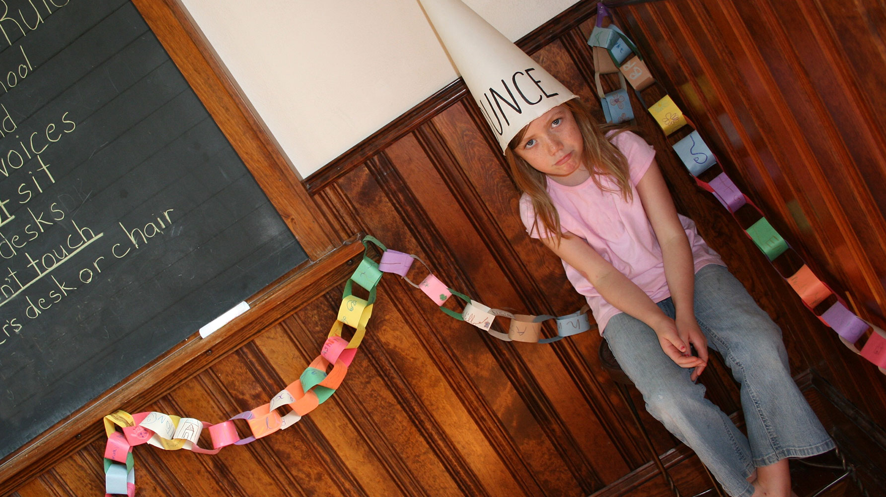 En genrebild av en flicka som sitter i en stol bredvid svarta tavlan, med en dumstrut på huvudet