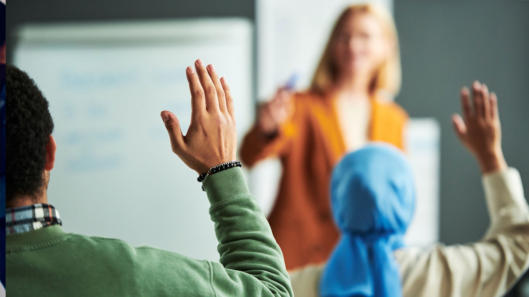 En genrebild av en lektion, läraren i oskärpa vid tavlan, en flicka i slöja räcker upp handen, eleverna är fotograferade bakifrån