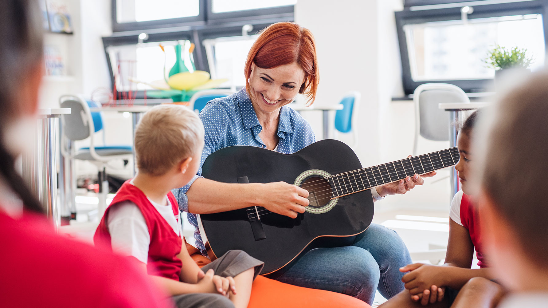 En genrebild av en lärare som sitter på golvet med en grupp barn, läraren spelar gitarr och har ögonkontakt med ett av barnen. Hon ser glad ut