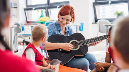 En genrebild av en lärare som sitter på golvet med en grupp barn, läraren spelar gitarr och har ögonkontakt med ett av barnen. Hon ser glad ut