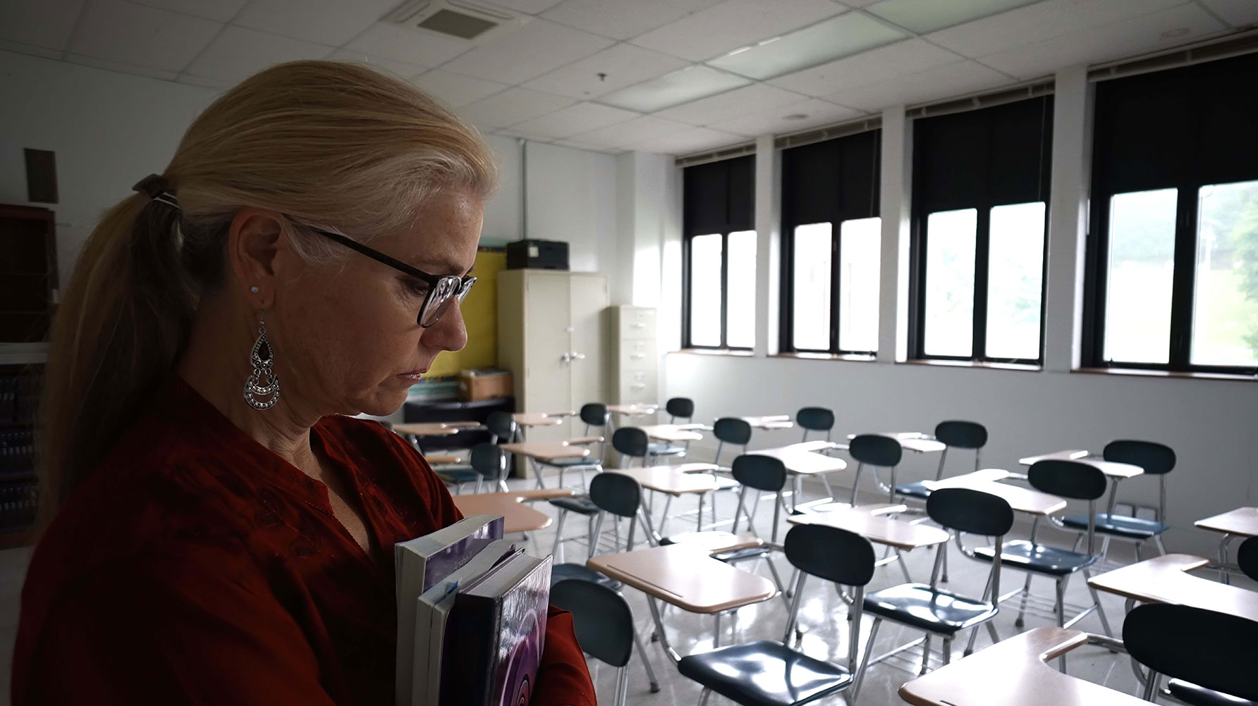 En genrebild av en kvinna i ett klassrum, hon ser allvarlig ut, bär böcker i famnen