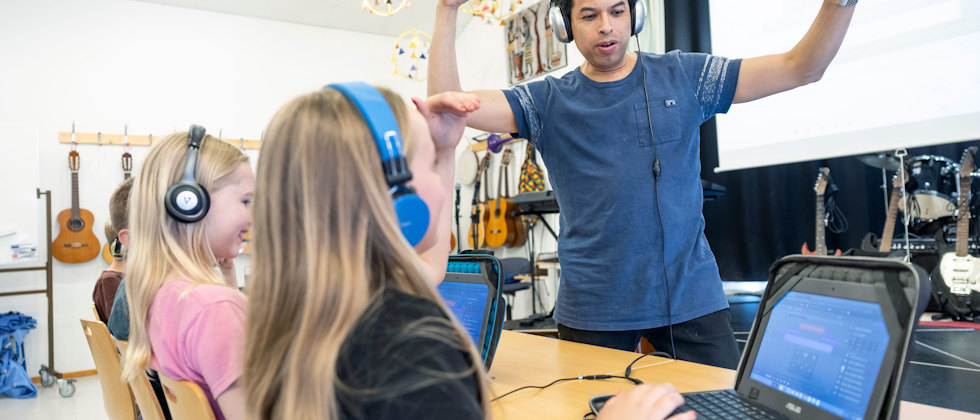 Musikläraren Martin Laps dansar med hörlurar i musiksalen medan tre elever sitter framför sina datorer och jobbar.