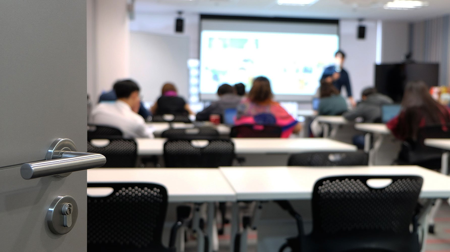 En genrebild av en d&#xF6;rr som st&#xE5;r &#xF6;ppen mot ett klassrum, lektionen p&#xE5;g&#xE5;r, fotograferad i osk&#xE4;rpa, l&#xE4;raren framme vid whiteboarden