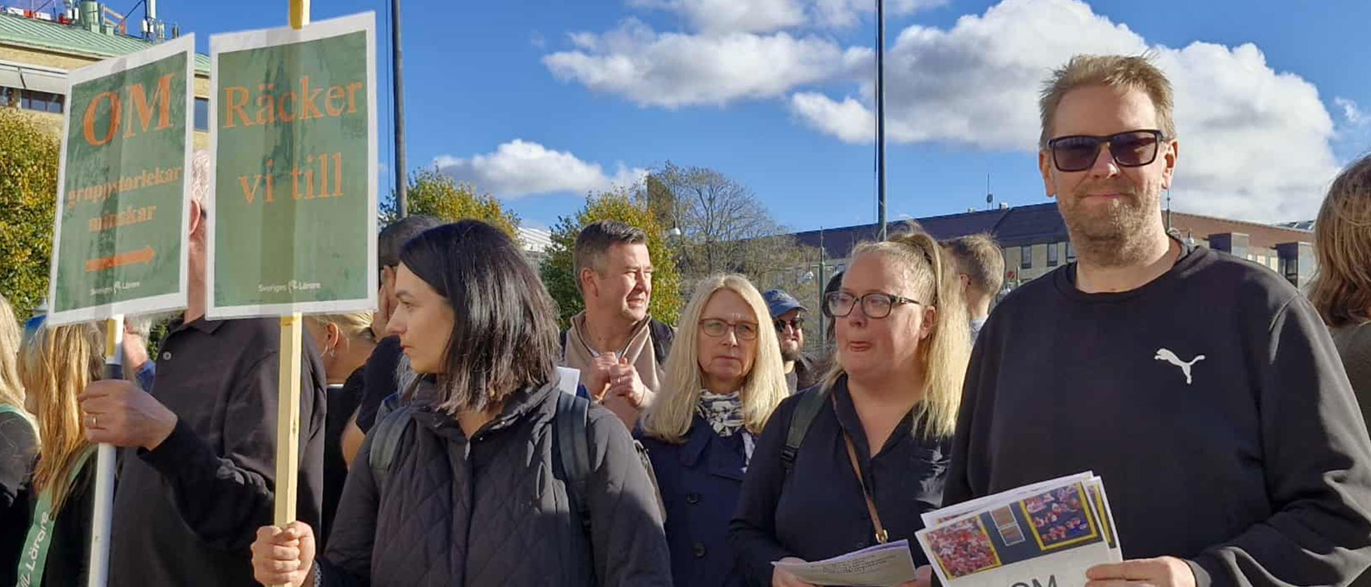 Lärare med skyltar med slagordet ”om” samlade på ett torg med blå himmel i bakgrunden