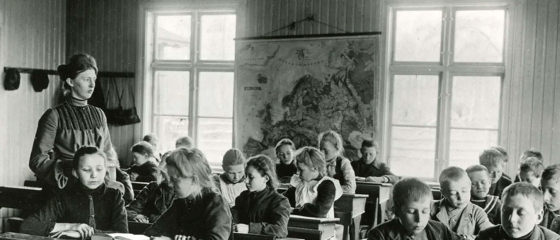 En svartvit bild av en klass kring förra sekelskiftet, en ung lärarinna med uppsatt hår till vänster i bild, koncentrerade barn klädda i sina ytterkläder i bänkarna