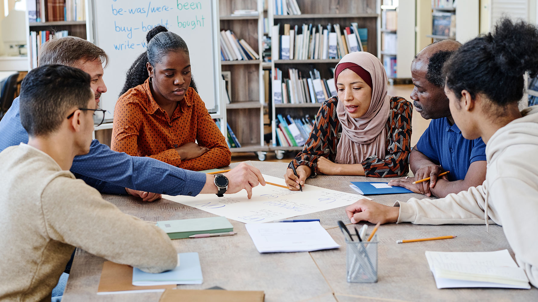En genrebild av en grupp människor runt ett bord, en bär hijab, de tittar på böckerna och papprena på bordet framför sig och verkar diskutera något
