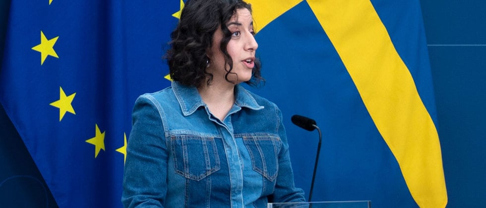 Ett foto av Simona Mohamsson talar i mikrofon framför EU:s flagga och den svenska flaggan vid en presskonferens