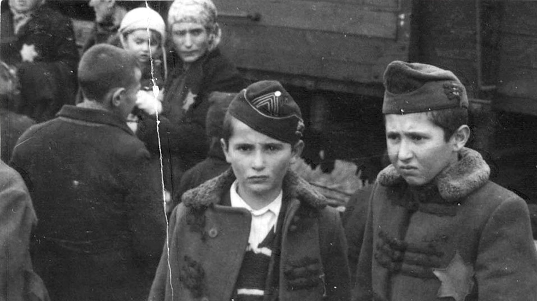 Ett svartvitt fotografi av två pojkar i kappa, i bakgrunden syns ett tåg, till vänster i bild står en grupp människor. En har en sexuddig stjärna fäst på bröstet.