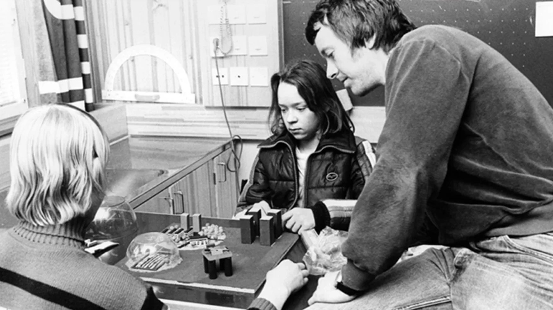 En svartvit bild av en manlig lärare som sitter på en skolbänk och undervisar två elever, av kläderna att döma sent sjuttiotal eller tidigt åttiotal